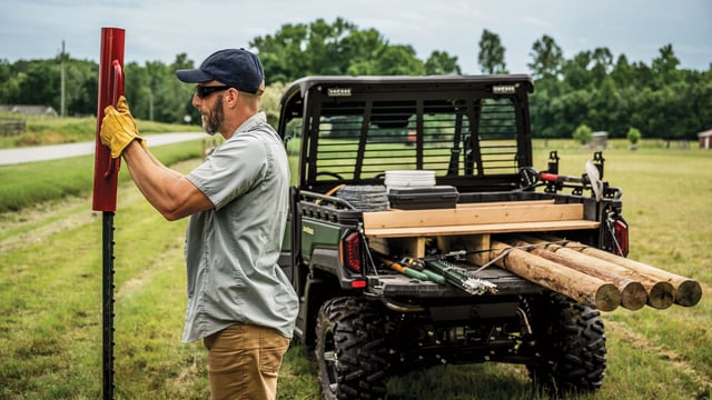 Man setting a T-post beside a John Deere Gator XUV with a cargo box loaded with fence posts, lumber and wire
