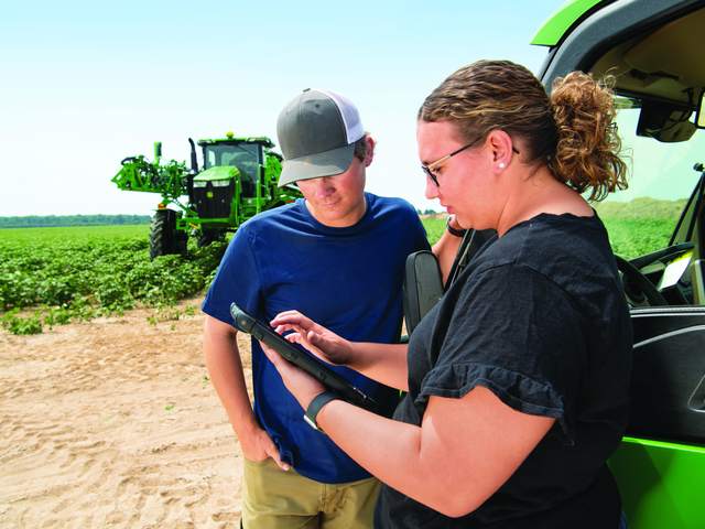 A farmer and a specialist reviewing a tablet beside a John Deere sprayer in the field